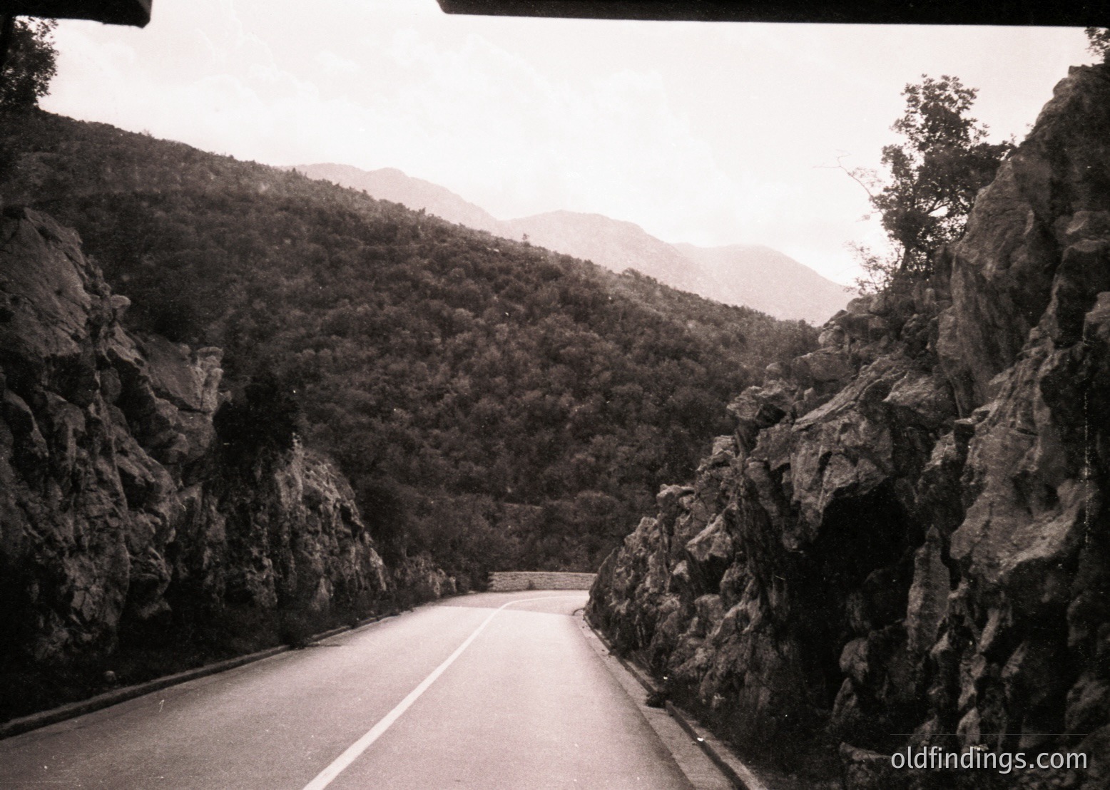Black-and-white shot of a winding mountain road flanked by steep, rocky cliffs and dense forest. Mid-20th century vintage, likely European alpine region. Asphalt road curves into misty distance, emphasizing isolation and rugged terrain.