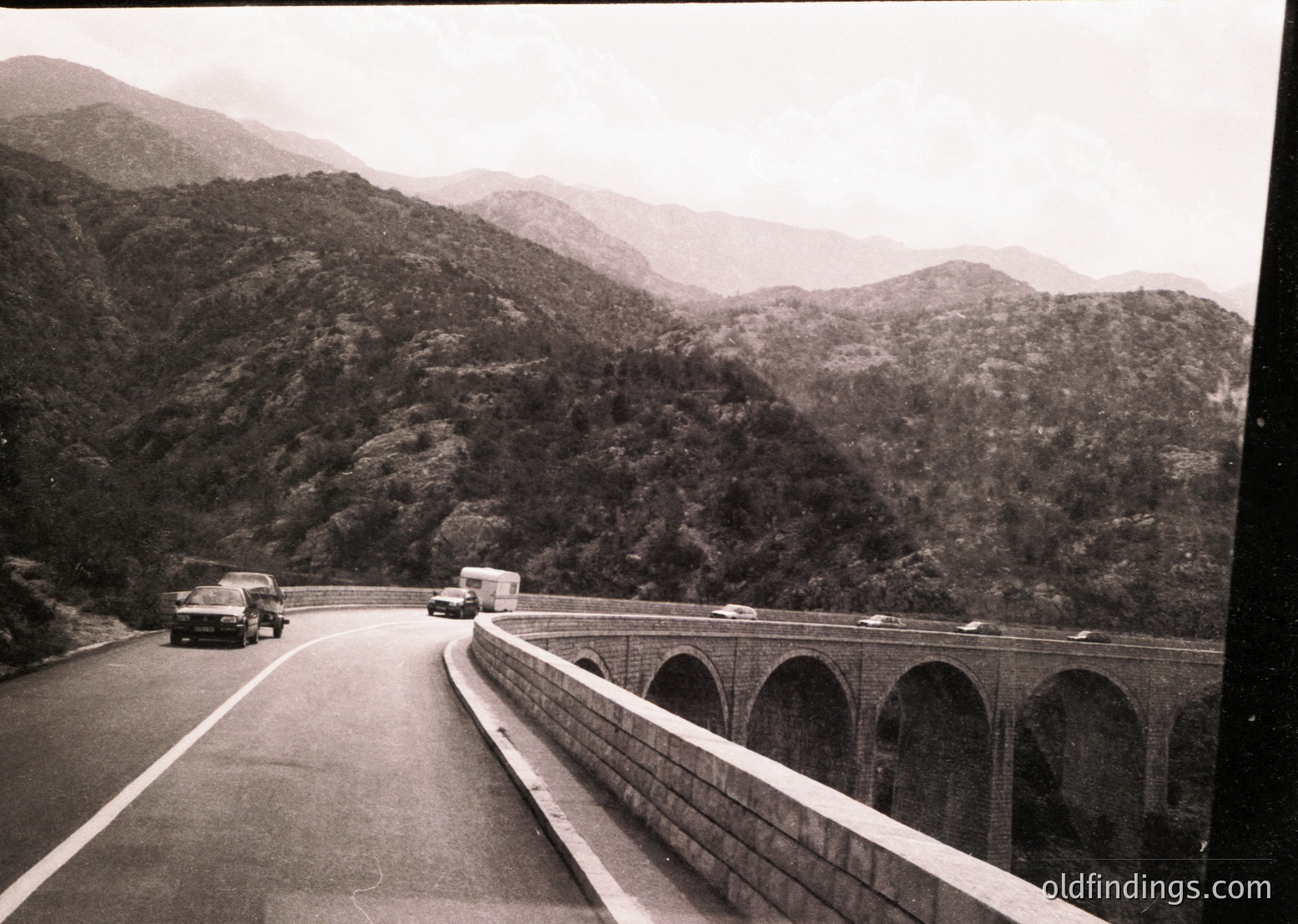 Mid-20th century highway bridge spanning lush, forested mountains. Concrete viaduct supports multiple lanes with vintage cars—trucks and sedans—traveling in both directions. Overcast skies enhance misty mountain backdrop.