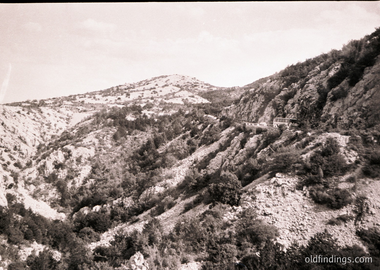 Black-and-white mountain landscape with rugged terrain and sparse vegetation, likely captured mid-20th century. Snow patches remain on higher elevations, contrasting with lower slopes. A narrow road winds upward, bordered by a simple guardrail. Overcast skies enhance the dramatic, timeless atmosphere.
