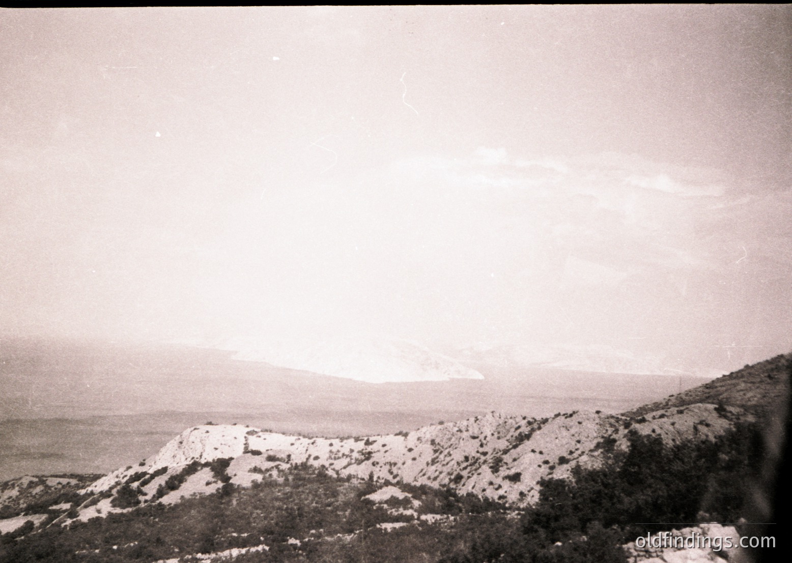 Vintage black-and-white seascape featuring rugged coastline with rocky cliffs and sparse vegetation. Distant shoreline meets calm waters under overcast skies, evoking mid-20th century coastal photography.