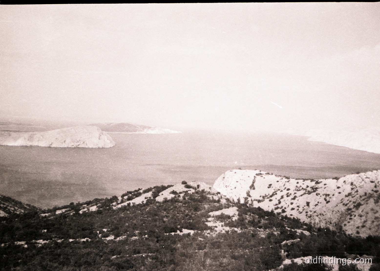 Vintage black-and-white coastal landscape featuring rugged cliffs and sparse vegetation overlooking a calm sea. Distant rocky outcrop and misty horizon suggest early 20th-century photography.