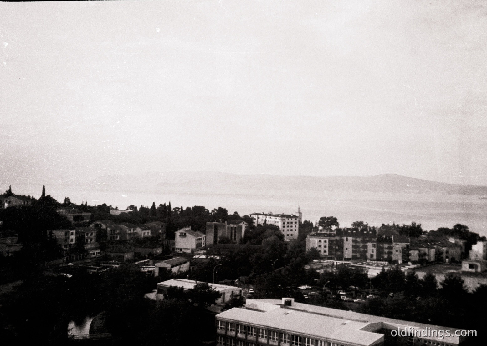 Black-and-white coastal cityscape featuring dense mid-20th-century architecture. Prominent multi-story buildings with flat roofs and minimalist designs line a hilly terrain overlooking a calm sea. A mosque with a tall minaret stands centrally, suggesting a Mediterranean or Balkan influence. Dense greenery separates residential blocks, hinting at urban planning prioritizing green spaces. Likely – era.