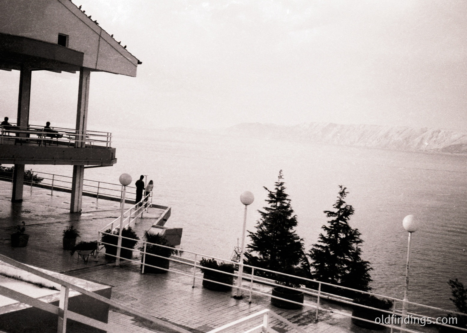 Mid-century seaside resort balcony overlooking misty waters, featuring brutalist concrete architecture with exposed pillars and railings. Two figures stand near spherical lampposts; evergreen trees frame the scene. Likely Eastern Bloc-era coastal design ( ).