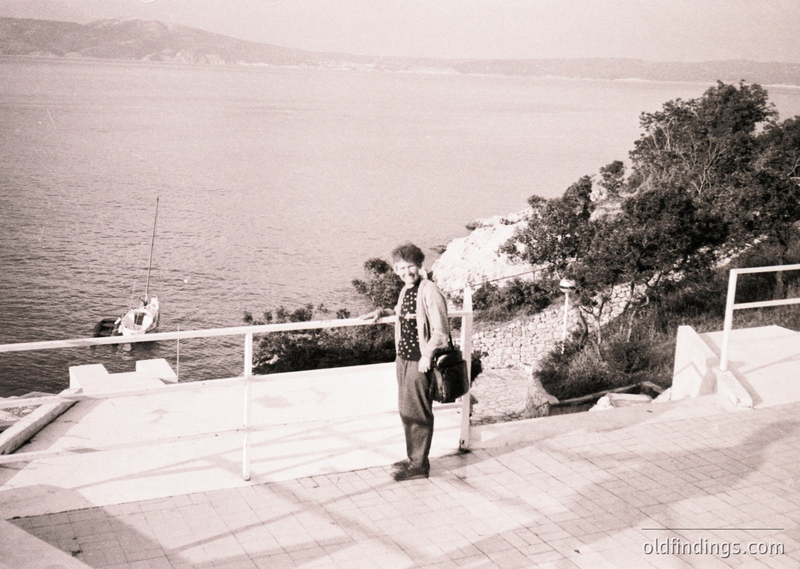 Mid-20th century seaside promenade with a lone woman in patterned dress and headscarf, holding a bag. White balustrade overlooks calm waters with a distant shoreline. Lush greenery and rocky terrain contrast with the paved walkway. Likely Mediterranean coastal town, 1950s-1960s.