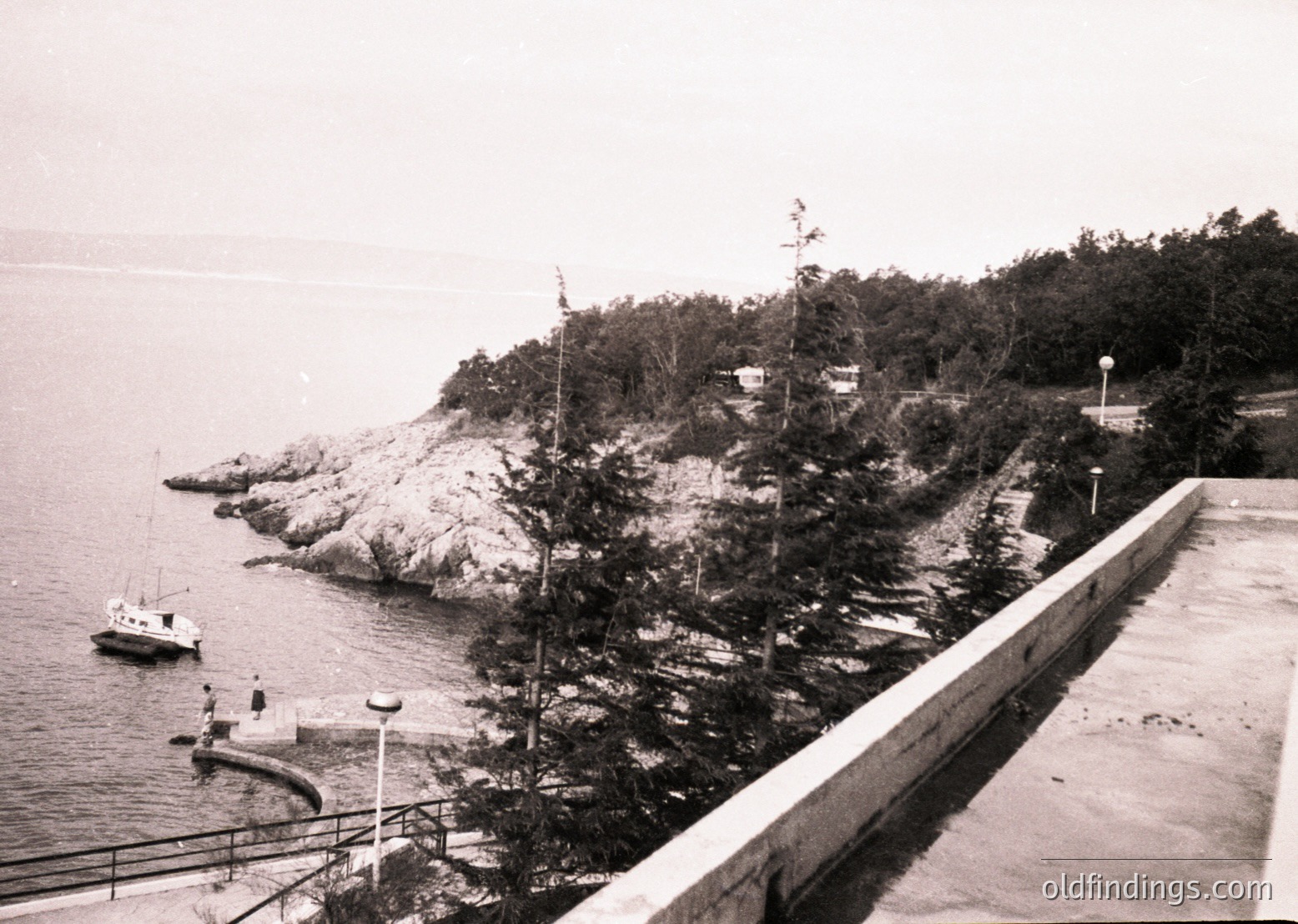 Black-and-white coastal scene featuring a concrete pier with railings extending into calm waters. Rugged cliffs with sparse vegetation and a lone boat anchored near shore. Mid-20th century architecture with minimalist lamp posts and a small structure atop the cliff.