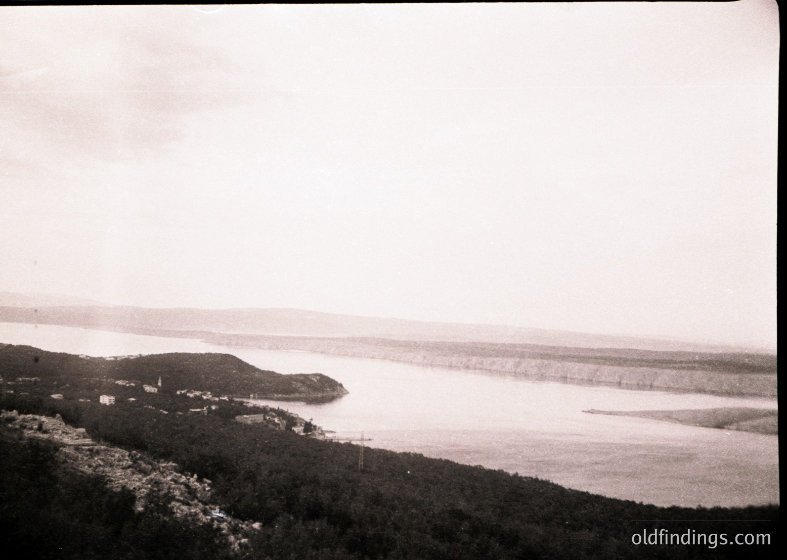 Black-and-white coastal landscape featuring a rocky shoreline and distant village by the sea. Dense greenery frames the left side, while a narrow strip of land extends into the horizon. Likely mid-20th century due to monochrome and composition style.