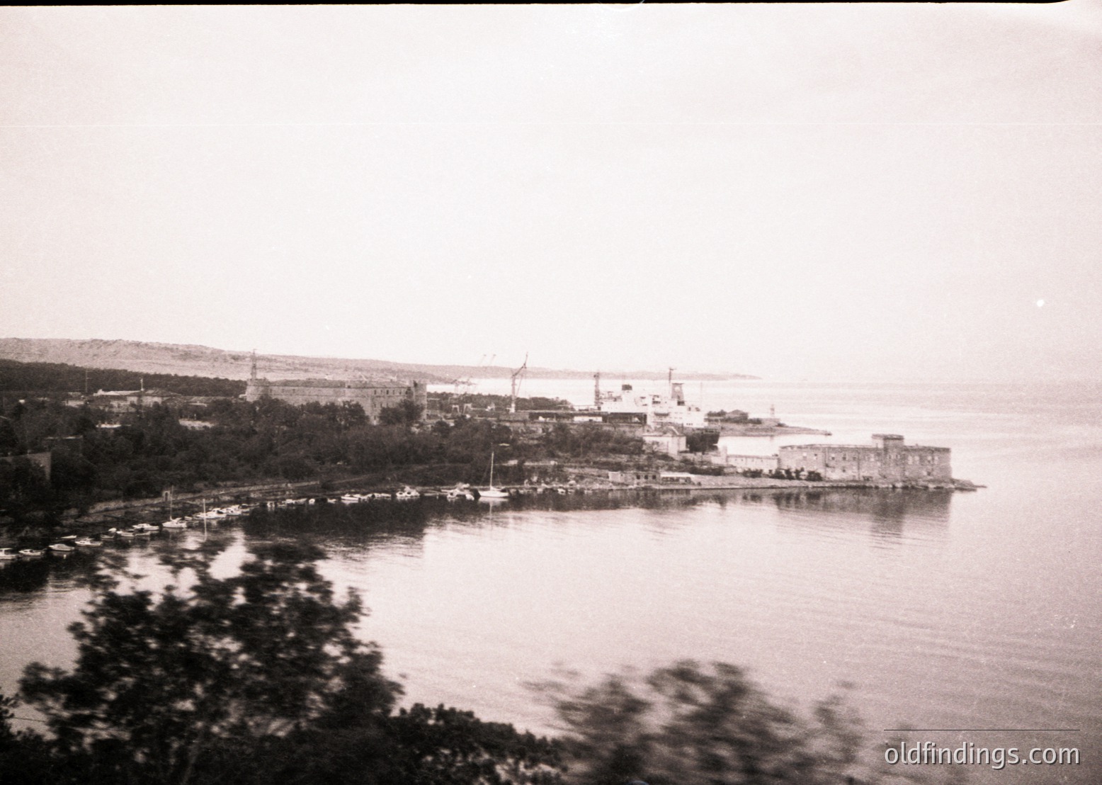 Black-and-white coastal industrial scene featuring a fortified stone structure near waterfront, likely a lighthouse or watchtower. Industrial buildings and cranes in background suggest port activity. Dense foliage frames left side. Vintage sepia tone indicates early-to-mid 20th century. Potential historical research value for maritime architecture.