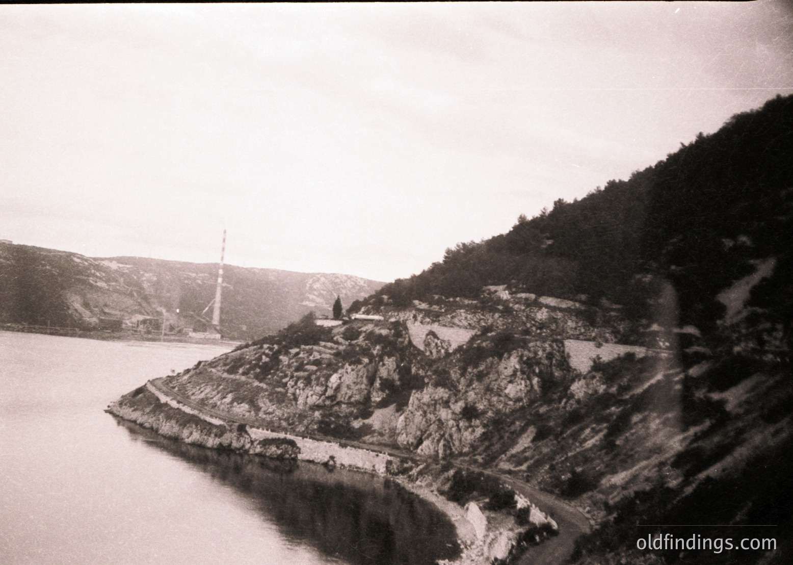 Vintage black-and-white coastal scene featuring a rocky promontory with a small, elevated structure—likely a lighthouse or chapel—perched on cliffside. Winding road hugs the shore, bordered by sparse vegetation and dramatic rock formations. Calm waters frame the left edge, with a mast or flagpole visible in the distance. Evokes early 20th-century European seaside architecture.