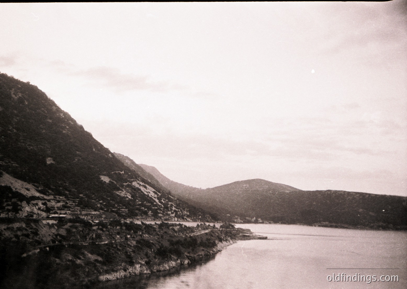 Vintage black-and-white lakeside scene with rugged mountain range framing a serene body of water. Dense forest clings to hillsides, meeting a winding shoreline. Likely early-to-mid 20th century European alpine region.