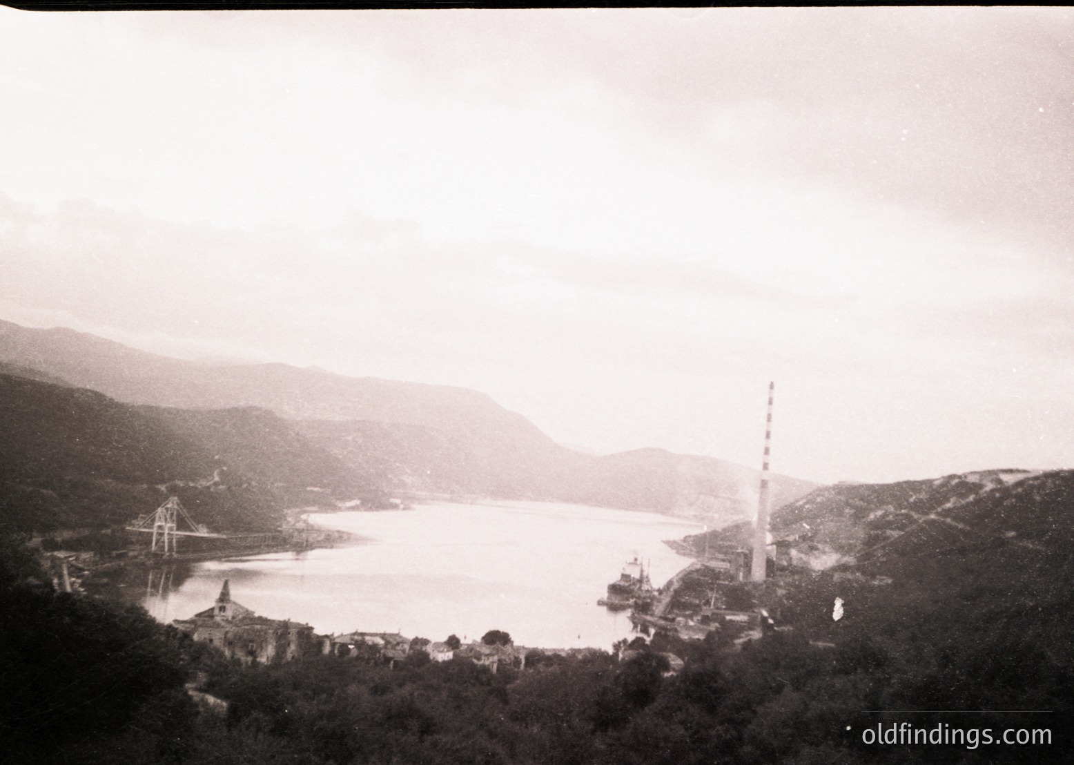 Vintage black-and-white coastal scene featuring a prominent radio tower on a hillside, a suspension bridge spanning a calm bay, and a small port town below. Mountainous terrain frames the horizon. Likely Eastern European, mid-20th century.
