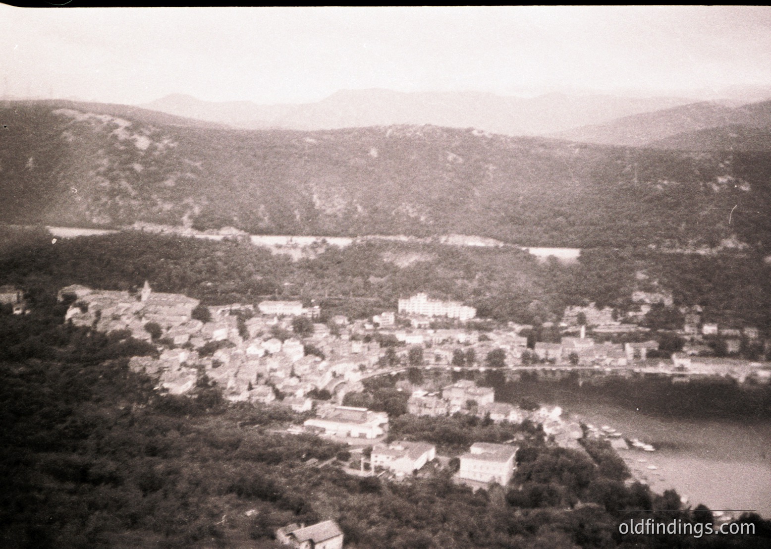 Aerial view of a coastal village nestled between dense greenery and a winding river, framed by mountainous terrain. Likely mid-20th century based on architectural style and monochrome tone.