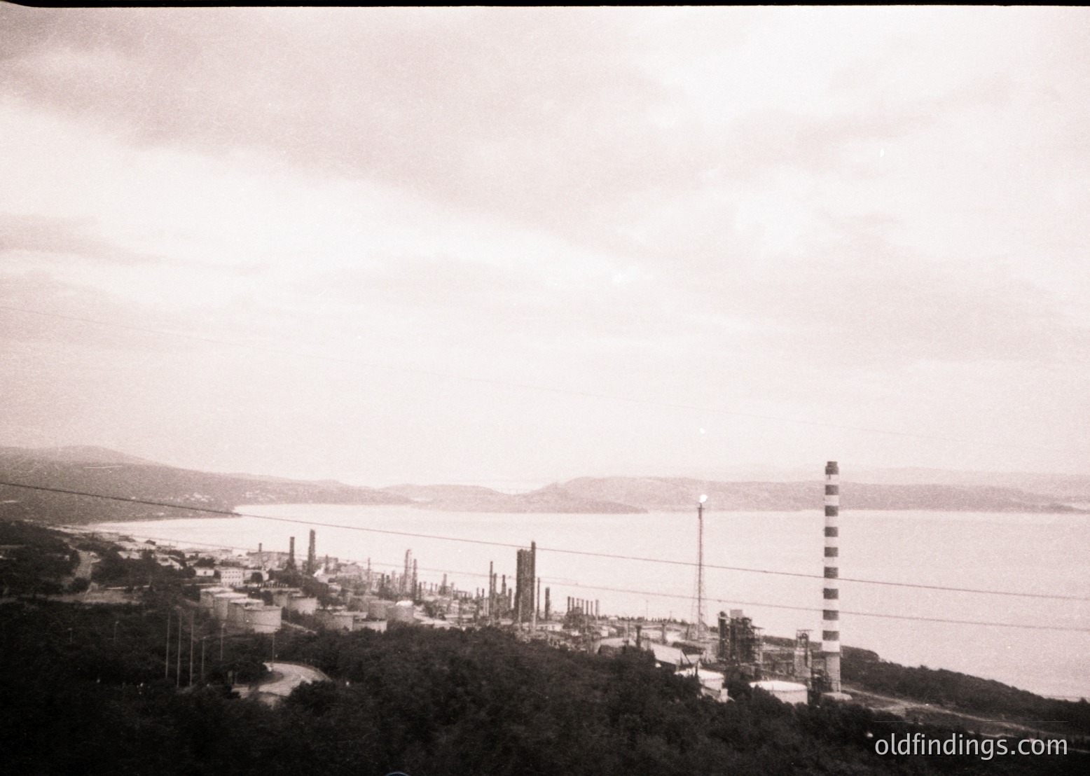 Black-and-white aerial view of an industrial coastal facility, likely a refinery or petrochemical plant, with multiple tall chimneys and storage tanks along a shoreline. Overcast skies and dense vegetation frame the scene, suggesting a mid-20th-century industrial landscape. Potential location: Eastern Europe or Middle East.
