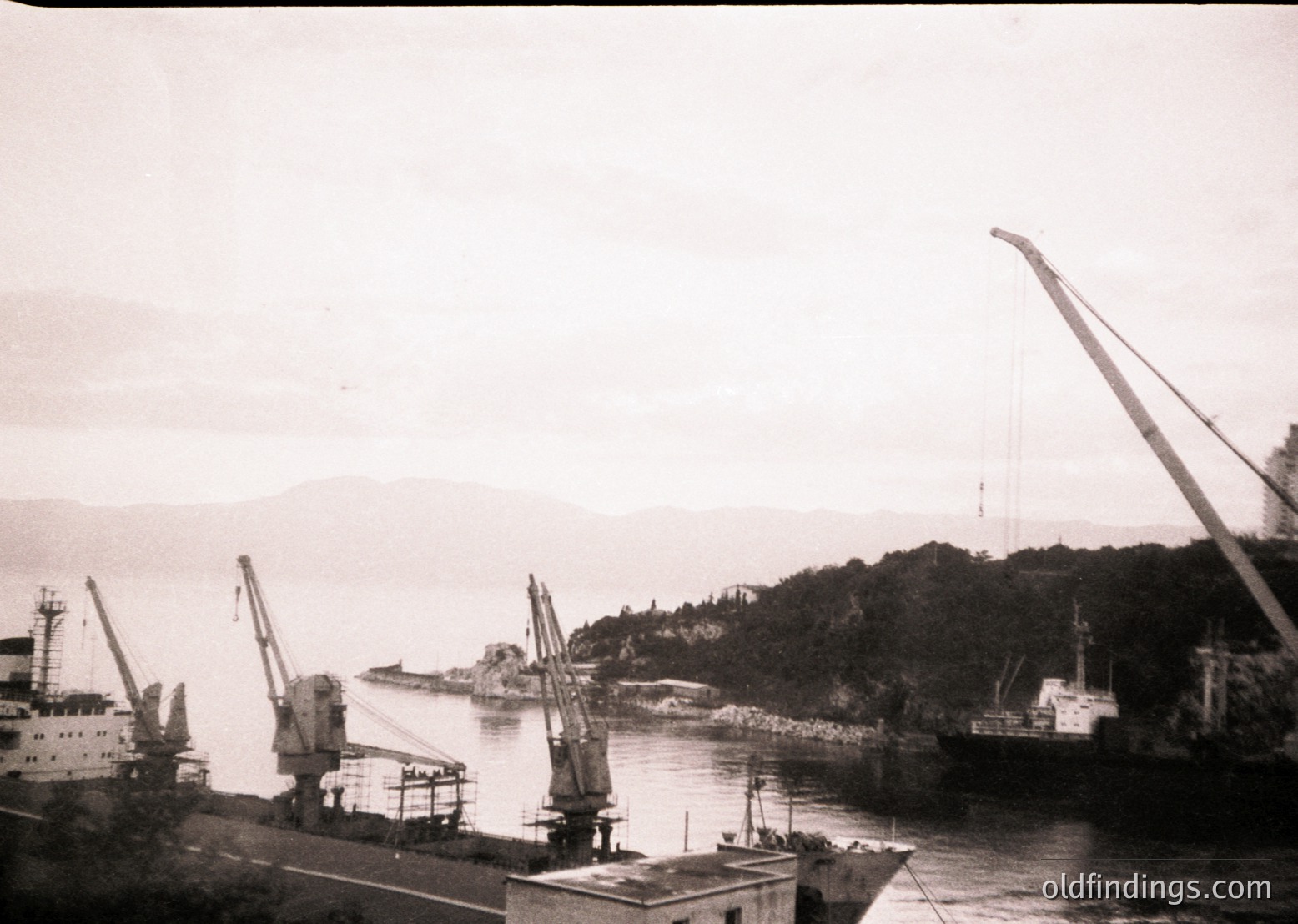 Black-and-white port scene featuring industrial cranes and docked vessels, likely mid-20th century. Coastal landscape with hills in background suggests a sheltered harbor. Vessels appear utilitarian, possibly for cargo or fishing.