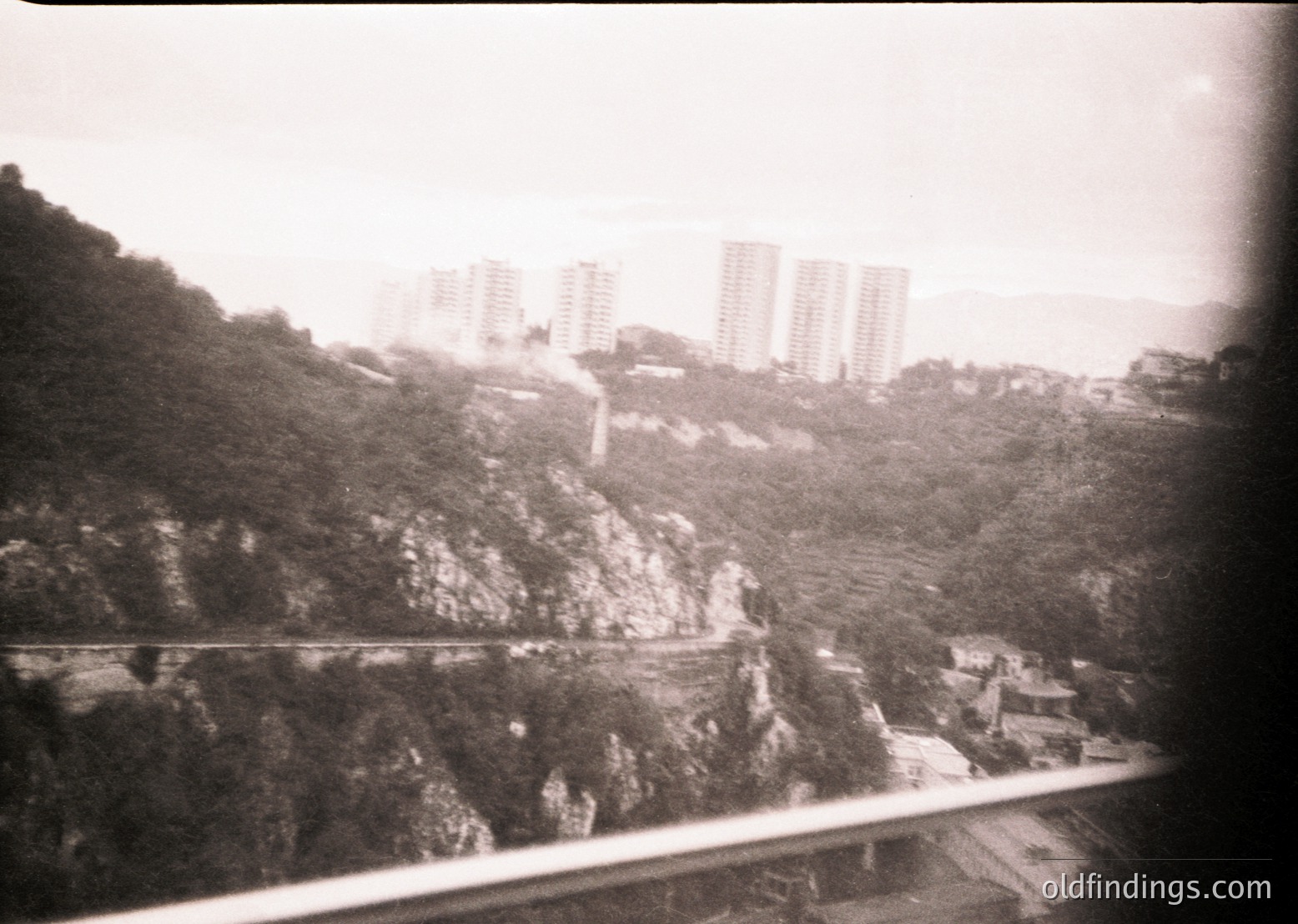 Vintage black-and-white urban landscape featuring high-rise apartment blocks on a hillside, likely from the mid-20th century. Curved road winding through rugged terrain below, framed by a window or train carriage. Evokes post-war Soviet-era architecture and urban planning.