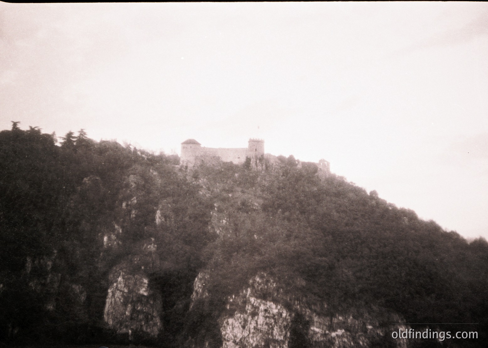 Medieval fortress perched atop rugged cliffs, surrounded by dense forest. Stone walls and towers suggest defensive architecture. Likely Eastern European, possibly or . Vintage sepia tone indicates or early century.