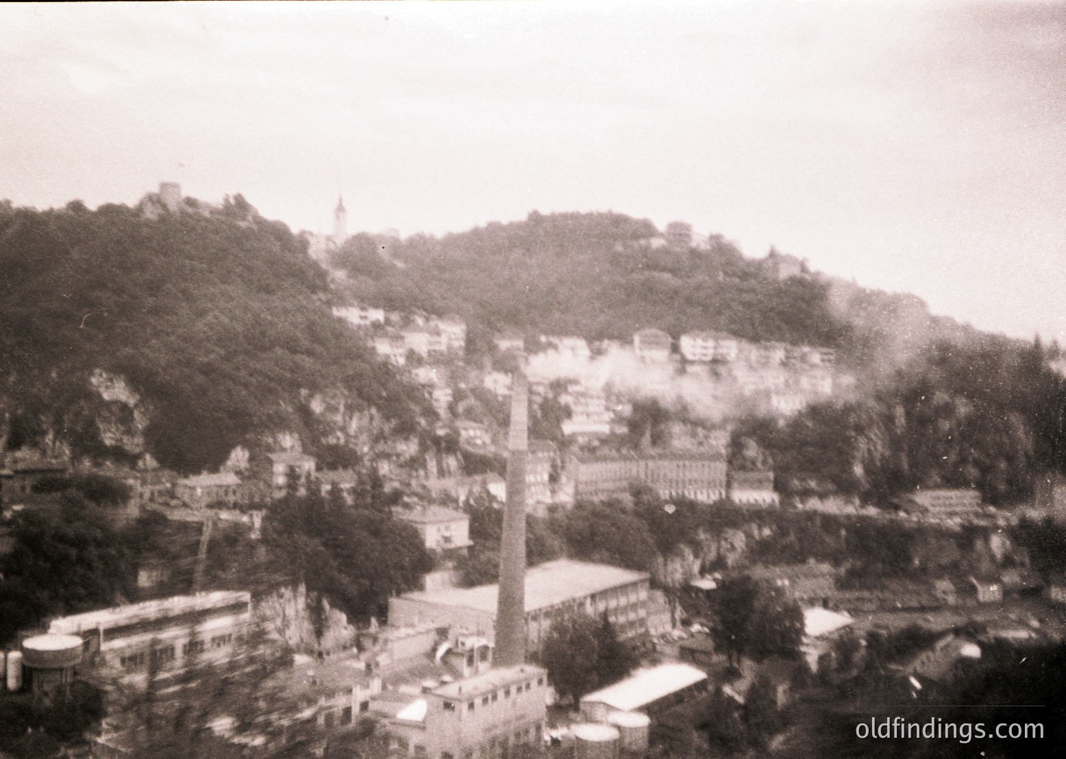Vintage black-and-white aerial view of a hilly coastal town with dense, irregularly built structures. Prominent industrial chimney and low-rise buildings dominate the foreground, while a castle-like fortress crowns the hillside. Dense greenery and rocky terrain frame the scene. Likely Eastern European, mid-20th century.