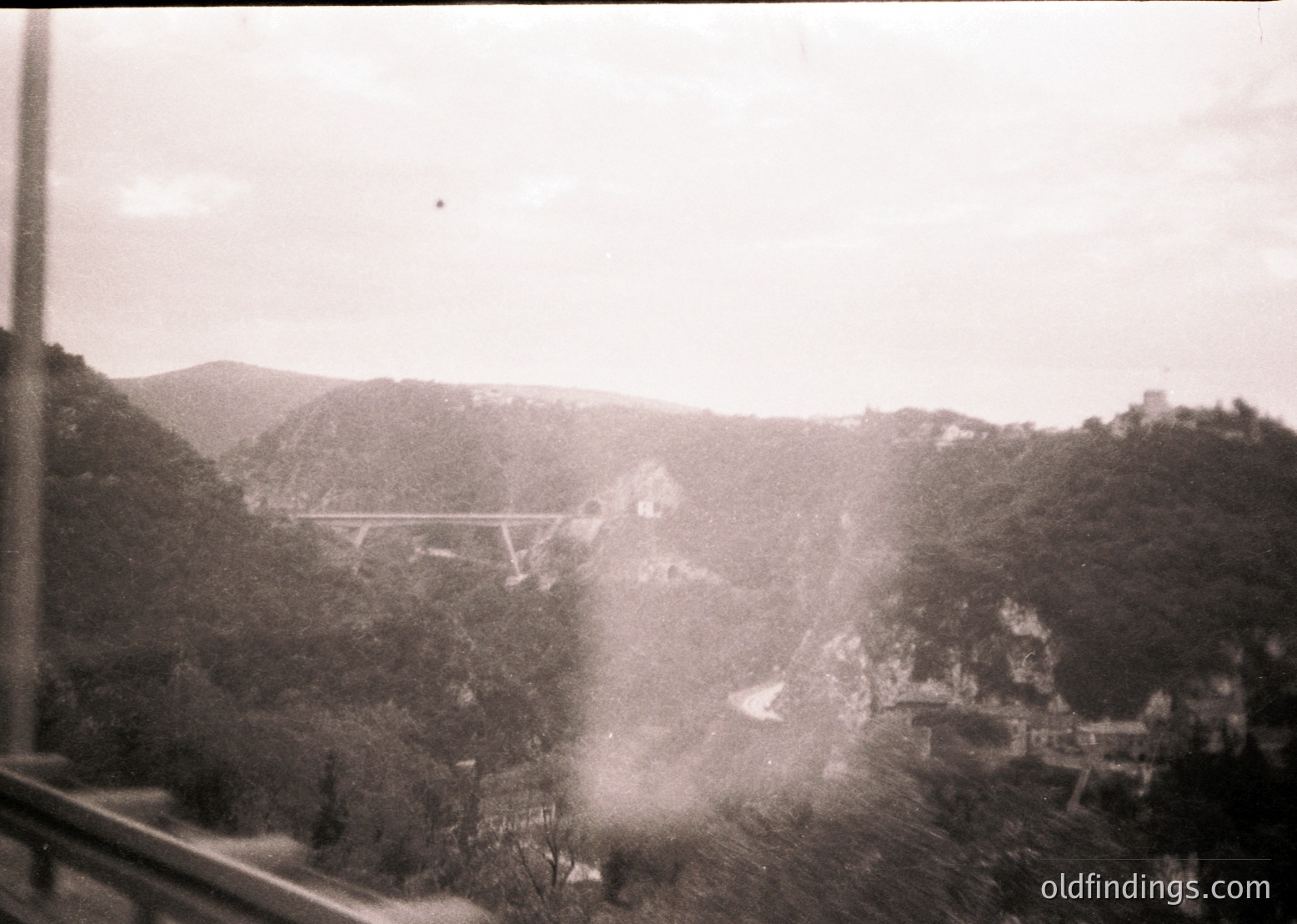 Vintage black-and-white view of a suspension bridge spanning a forested valley, framed by a window. Mid-20th century industrial or tourist architecture visible below. Dense foliage and misty terrain suggest mountainous terrain.