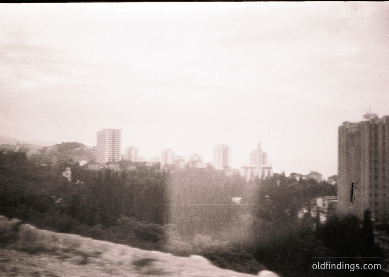 Urban skyline with mid-rise buildings and dense greenery, likely from a 1960s–1970s monochrome photograph. Fog or mist obscures distant structures, creating a hazy atmosphere. Architectural style suggests Soviet-era construction.