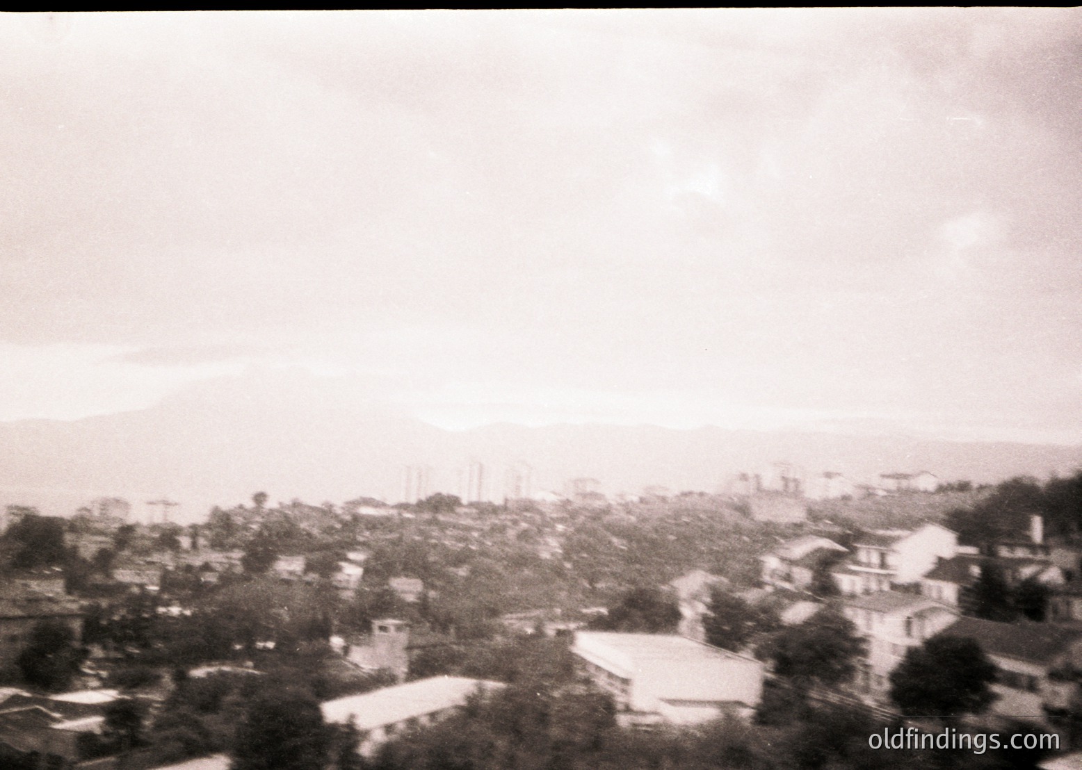 Vintage black-and-white aerial view of a densely built coastal town with low-rise buildings and scattered greenery. Distinctive industrial chimneys and a distant mountain range frame the horizon. Likely Mediterranean or Adriatic region, 1950s–1970s.