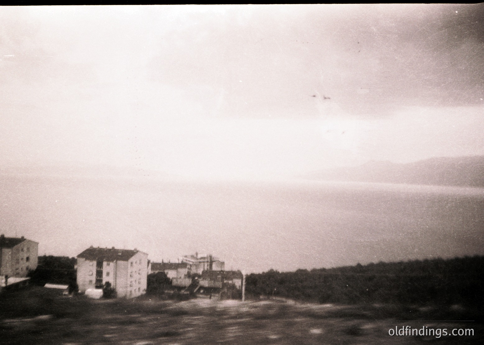 Mid-20th century seaside town with Soviet-era concrete buildings by the shore. Low-rise apartment blocks and a central structure dominate the flat landscape, framed by a misty horizon. Vintage black-and-white grain enhances nostalgic atmosphere. Likely Eastern Bloc coastal architecture, 1950s–1970s.