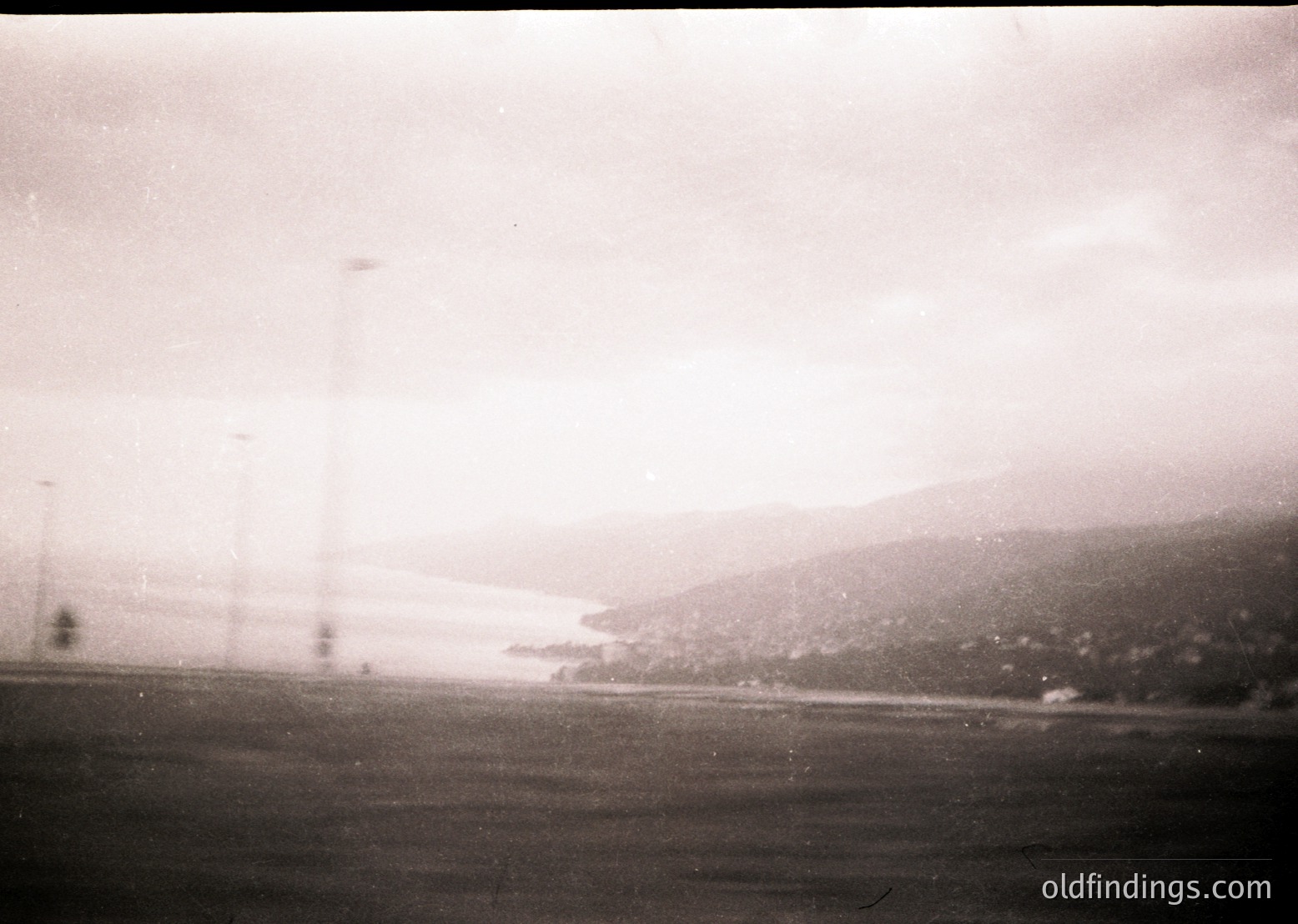 Vintage black-and-white shot of a rural landscape with power lines stretching into the distance, framing a hazy horizon. Low-lying hills and sparse vegetation suggest a flat terrain, possibly agricultural. Film grain and soft focus hint at mid-20th century photography.