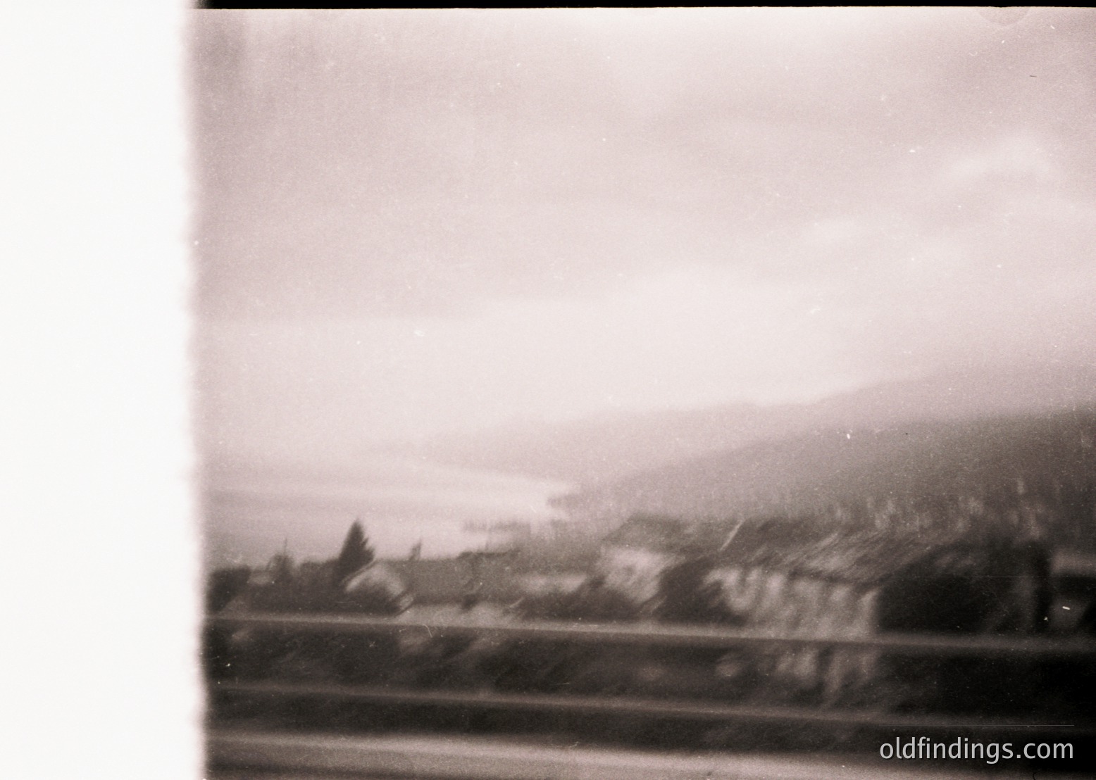 Vintage black-and-white train window view of rural landscape, featuring misty hills and scattered buildings. Likely mid-20th century due to grainy film texture.