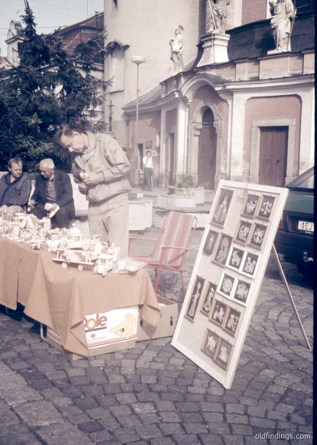 Street-side vintage market in a European town square, likely 1960s–70s. Three men examine ceramic figurines and framed religious icons displayed on a table draped with a beige cloth. Baroque-style church façade with statues and arched doorway in background. Cobblestone pavement and parked car suggest small-town charm.