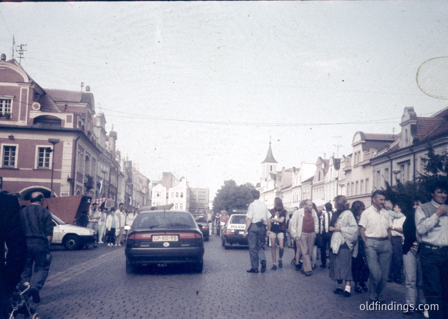Vintage street scene with cobblestone road, mid-20th century European architecture, and dense pedestrian traffic. A mix of vintage cars and pedestrians in casual 1960s–1970s attire. Prominent church spire in background suggests historic town center.