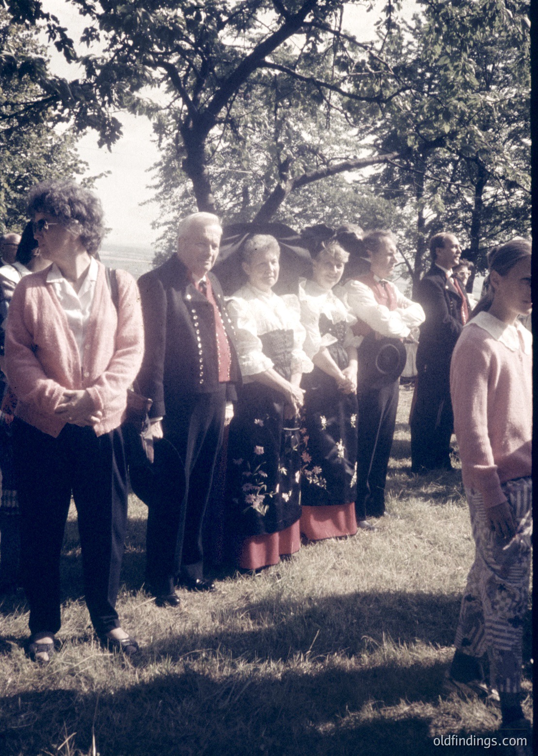 Group of 10+ individuals in outdoor ceremony, likely 1970s-1980s Eastern Bloc. Central figure in traditional embroidered dress and red sash, flanked by men in formal suits and women in light-colored blouses. Photographer visible on right, capturing event. Lush greenery and trees suggest rural or park setting.