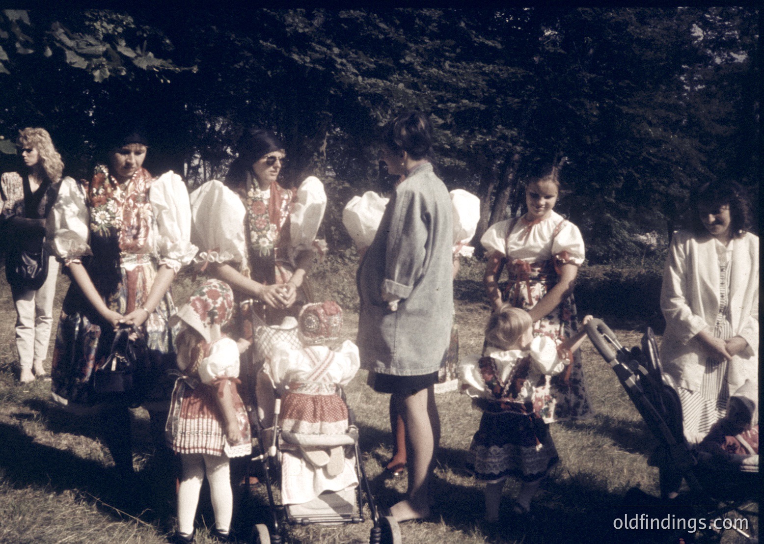 Vintage outdoor gathering of women and children in folk-inspired attire, likely Eastern European, mid-20th century. Women wear embroidered blouses and headscarves; children in simple dresses and aprons. One woman holds a child in a woven basket, others interact with strollers. Lush greenery and trees frame the scene, suggesting a rural or village setting.