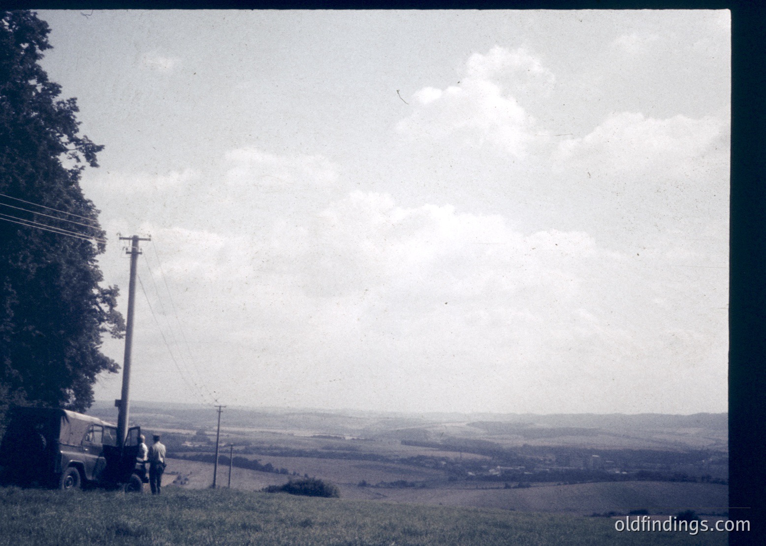 Vintage black-and-white rural landscape featuring a lone utility pole and two figures near a vintage truck on a grassy hillside. Open fields stretch toward distant treeline under a cloudy sky. Mid-20th century farming or travel scene.
