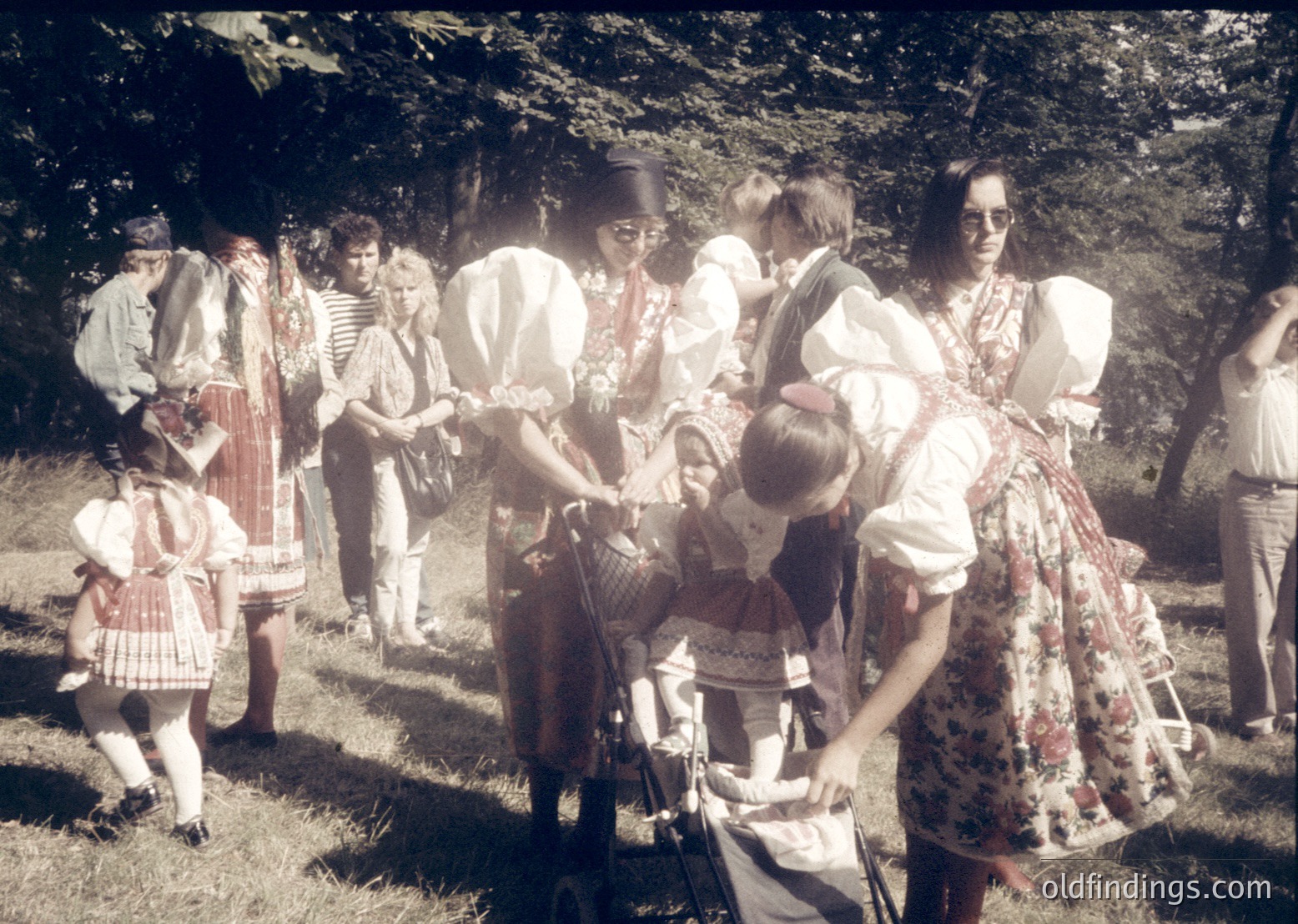 Group of adults and children in traditional folk attire, likely Eastern European, gathered outdoors in a rural setting. Men wear embroidered vests and hats; women in layered blouses and floral aprons. A child in a basket on someone’s back suggests a communal, possibly agricultural or festival scene. Warm lighting and greenery hint at summer.