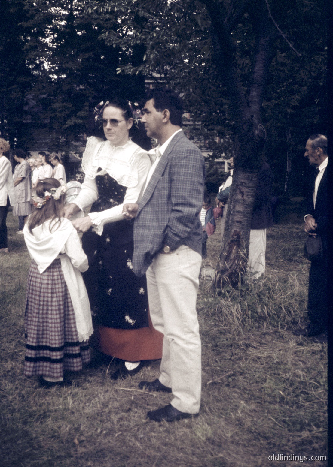 Mid-20th century outdoor gathering with formal attire: man in checkered blazer, white trousers, and sunglasses shaking hands with a woman in a floral dress and hat. A young girl in a white dress with plaid skirt stands beside her. Wooden fence and trees frame the scene, suggesting a rural or park setting.