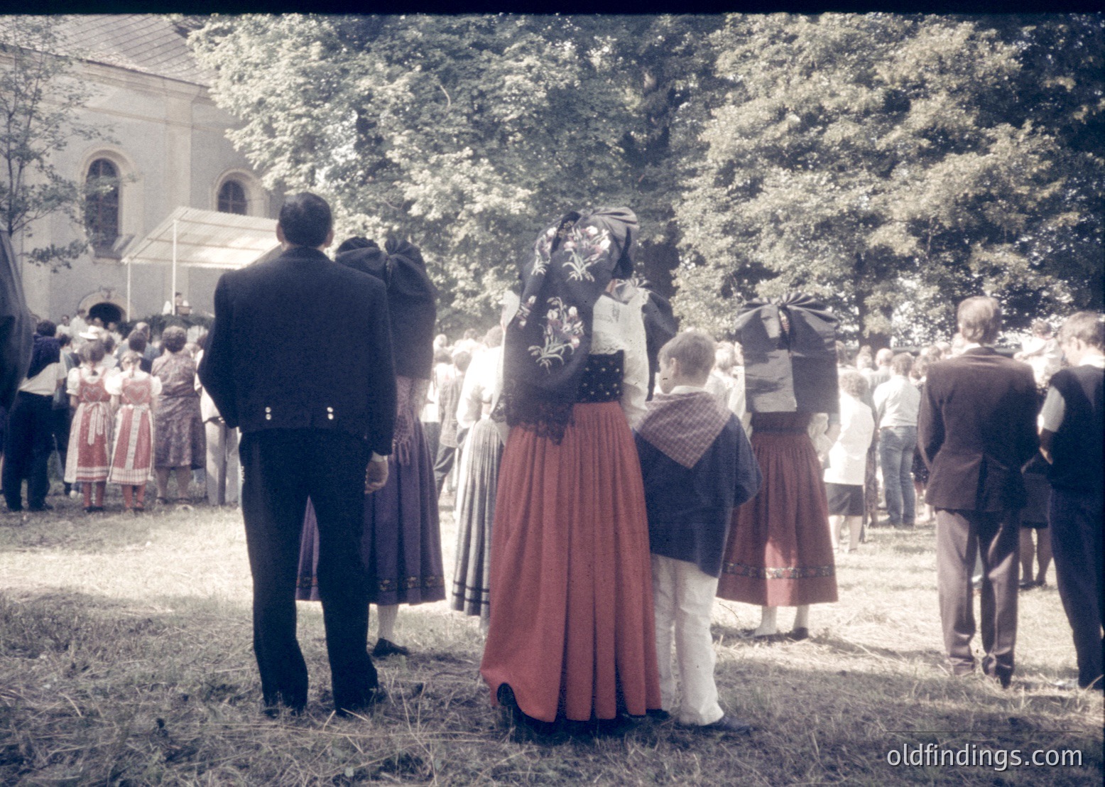 Vintage outdoor gathering featuring traditional folk attire—men in dark suits, women in embroidered skirts and blouses with floral headscarves. Crowd faces a white church with arched windows under mature trees. Likely Eastern European cultural festival, 1960s–1980s.