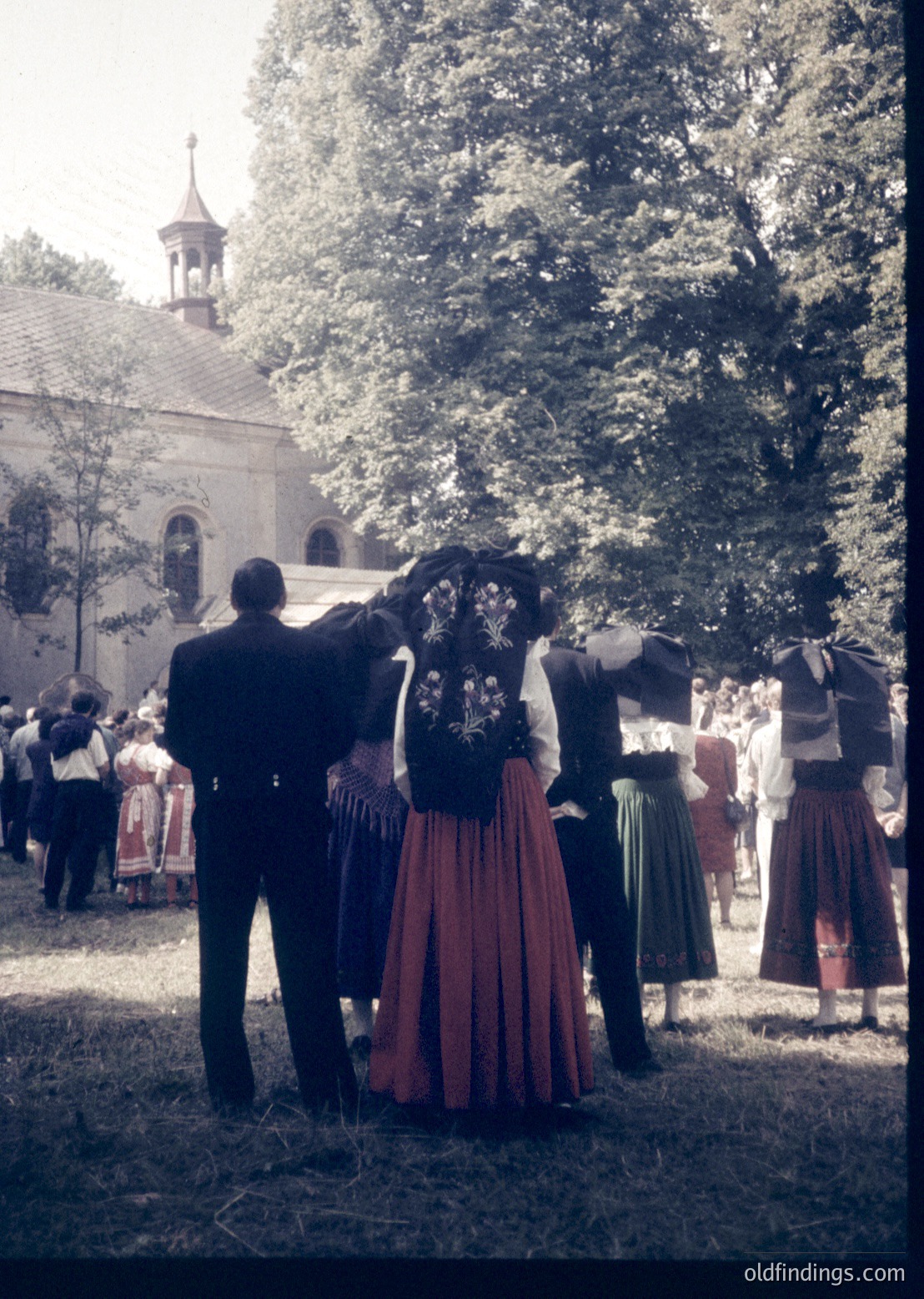 Vintage outdoor gathering featuring traditional folk attire—men in dark suits with white shirts, women in embroidered dresses with red skirts and green aprons. A church with a spire and arched windows forms the backdrop. Likely Eastern European cultural festival, mid-20th century.