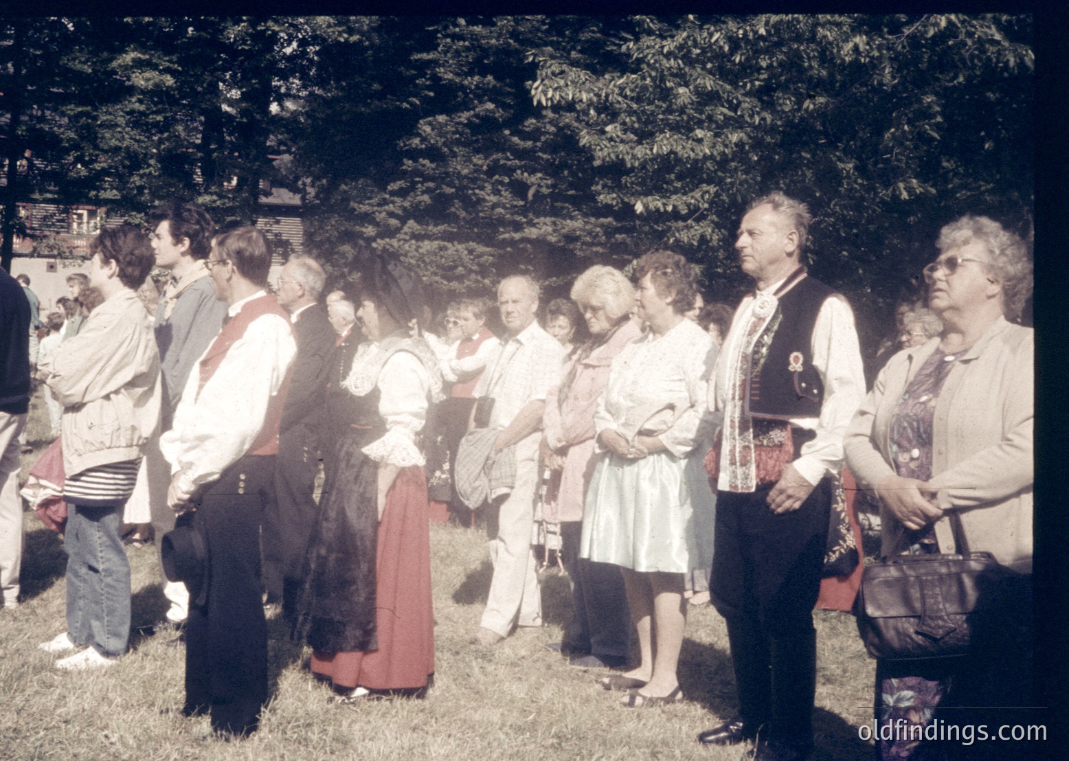 Group of adults in traditional folkwear, likely Eastern European, gathered outdoors in a wooded area. Men wear embroidered vests and shirts, women in long skirts and blouses. Mid-20th century (1950s–1970s) attire suggests cultural festival or heritage event. Lush greenery and partial brick wall in background.