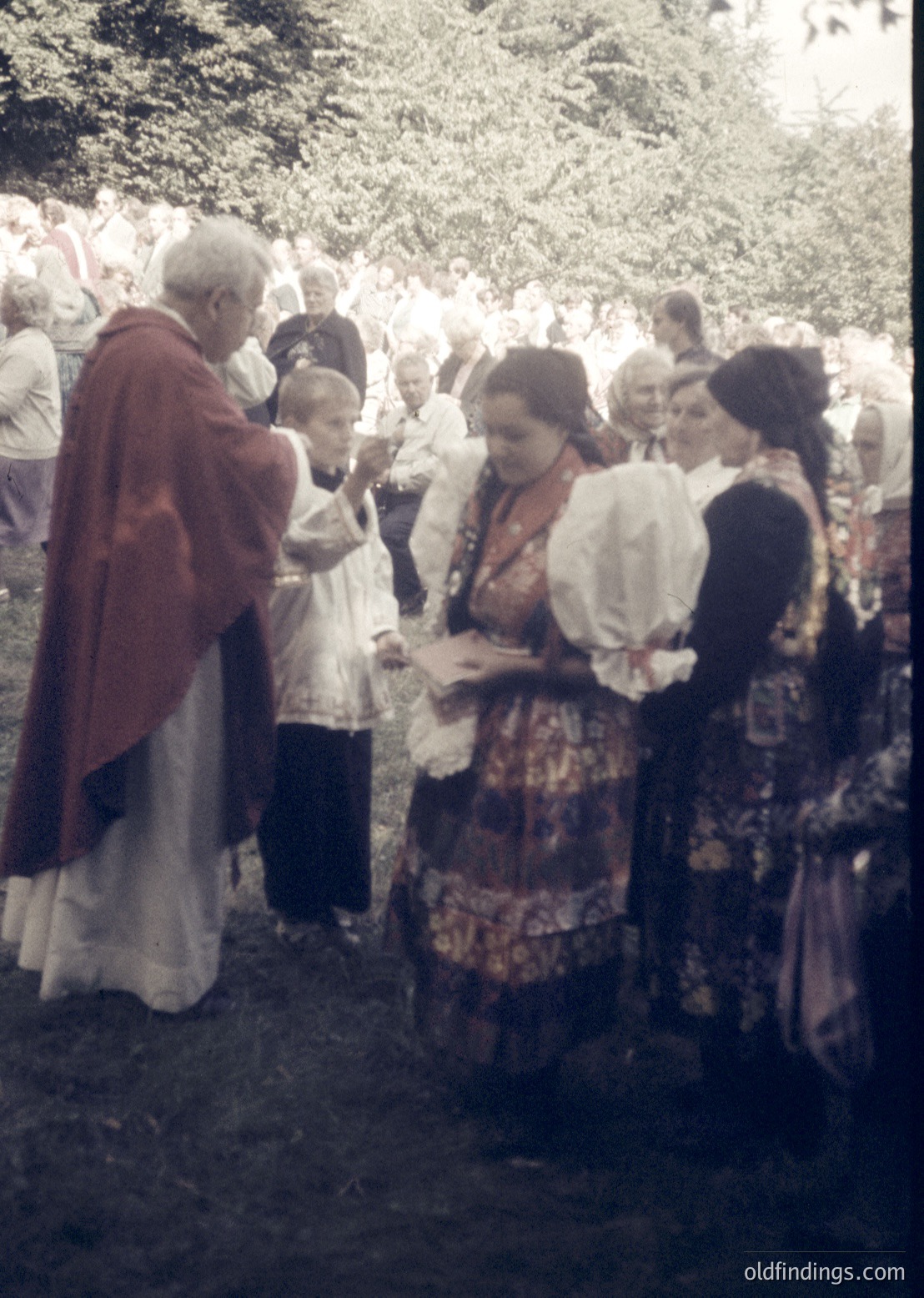Vintage outdoor gathering featuring a priest in liturgical vestments blessing a woman in traditional folk attire, surrounded by a crowd in rural attire. Likely Eastern European, mid-20th century (1950s–1960s).