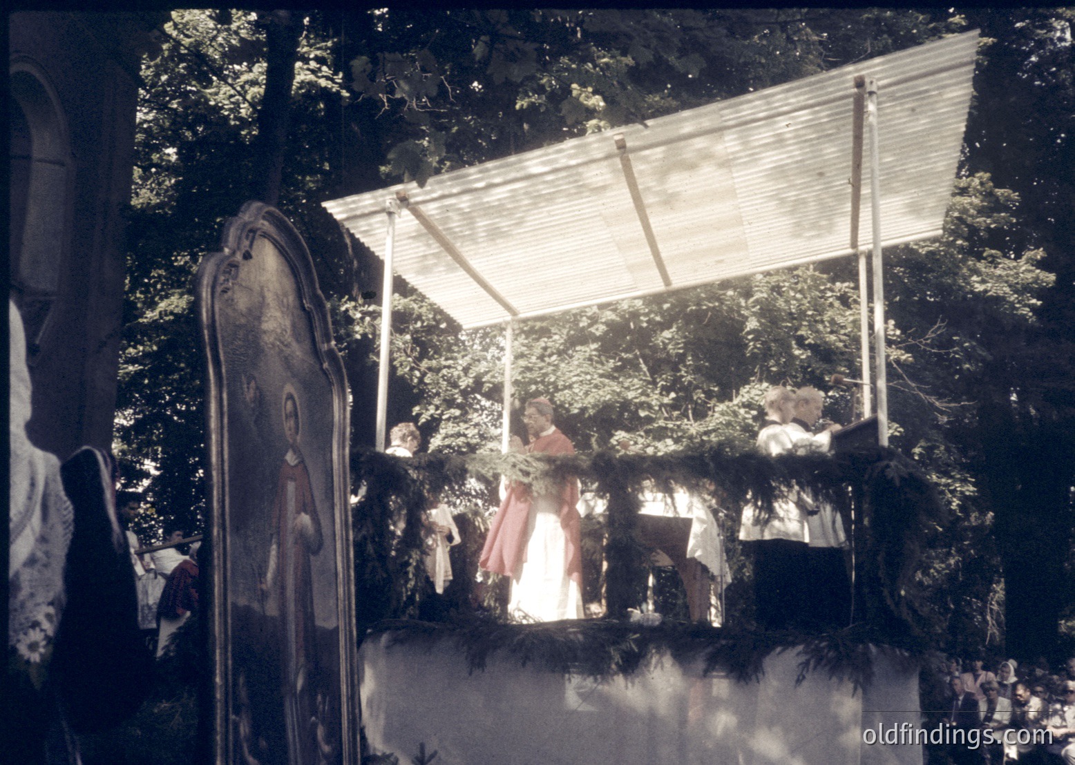 Vintage outdoor stage with wooden canopy and draped fabric, featuring a woman in a 1960s-style dress performing on a raised platform. Audience seated on wooden benches beneath leafy trees, suggesting a rural or folk festival setting.