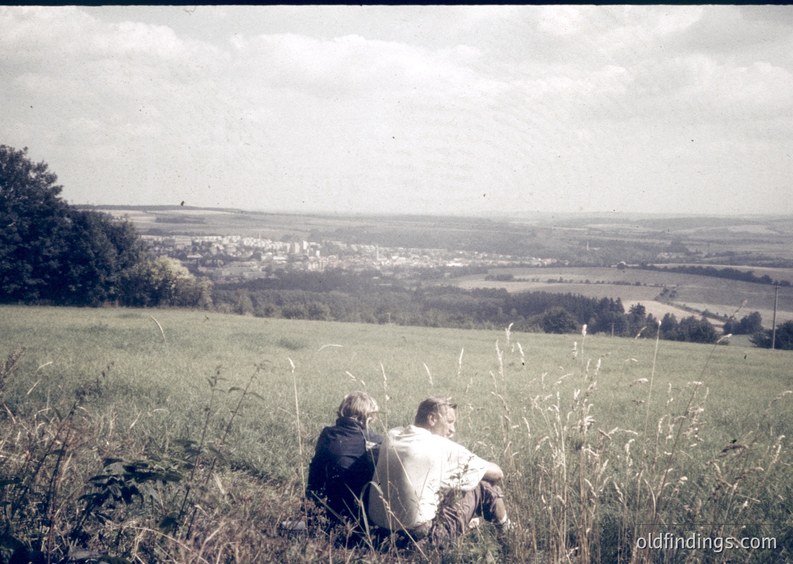 Two individuals sit on a grassy hillside overlooking a rural landscape, likely mid-20th century. The sepia-toned photo captures a serene countryside scene with scattered trees, fields, and a distant village.