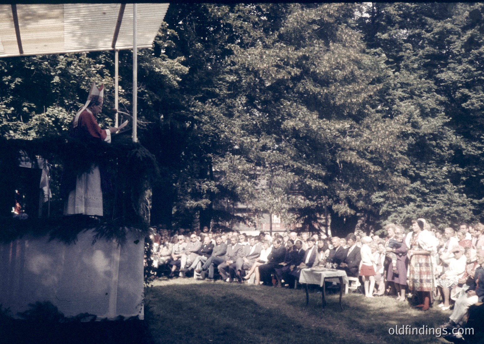 Outdoor gathering under dense trees with a stage on left. Speaker on elevated platform addresses seated audience in traditional attire; vintage sepia tone suggests mid-20th century. Possible cultural festival or community event.
