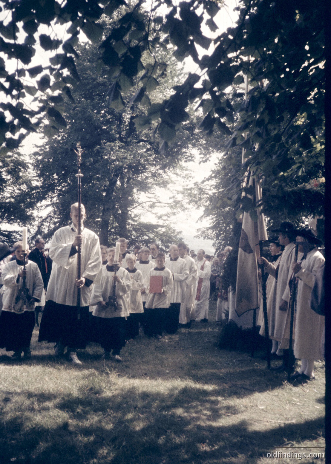Religious procession featuring clergy in white vestments, holding candles and banners under dappled sunlight through trees. Likely Eastern Orthodox given vestments and ritual objects. Outdoor setting suggests a rural or park-like location. Style and attire suggest mid-20th century (1950s–1970s).