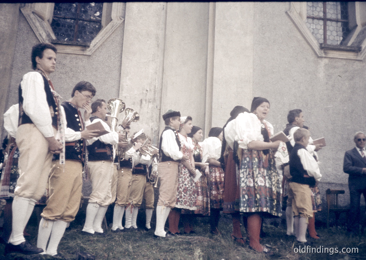 Traditional folk ensemble performing outdoors, likely Eastern European, mid-20th century. Men in embroidered vests and white trousers play brass instruments; women in floral dresses and headscarves sing. Stone church facade in background suggests cultural festival or village celebration.