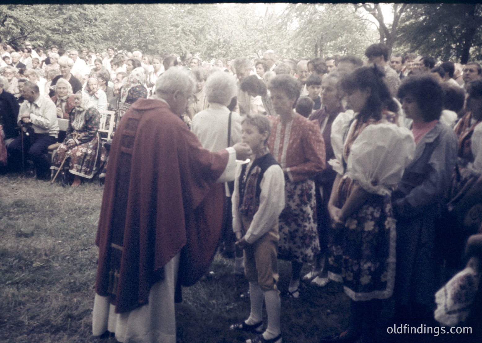 Outdoor religious ceremony with a priest in liturgical vestments blessing a young boy in traditional attire. Crowd of adults in folk costumes, seated on folding chairs under trees. Mid-20th century European rural setting.