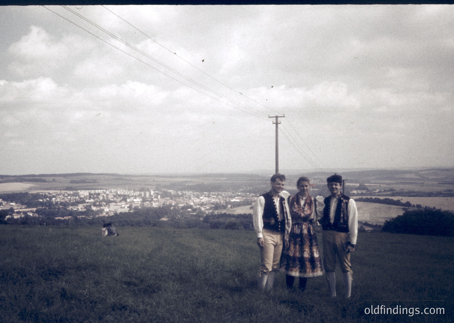 Three individuals in traditional folkwear pose on a grassy hilltop, overlooking a rural settlement. The man on the left wears a vest and cap, the woman a patterned dress, and the man on the right a vest with a headscarf. Sepia-toned vintage photo suggests mid-20th century Eastern European countryside.