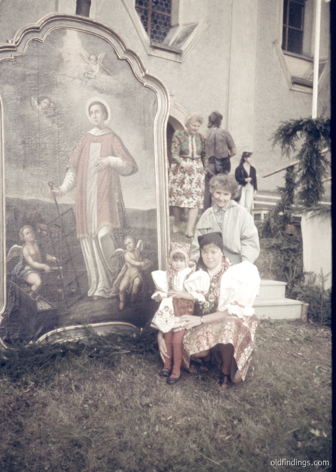 Vintage sepia-toned portrait of a woman and child seated on steps beside a religious mural depicting a saint flanked by cherubs. The woman wears traditional embroidered attire with a headscarf; the child holds a small box. Background shows a crowd near a building with arched windows, suggesting a public or ceremonial setting. Likely Eastern European, mid-20th century.