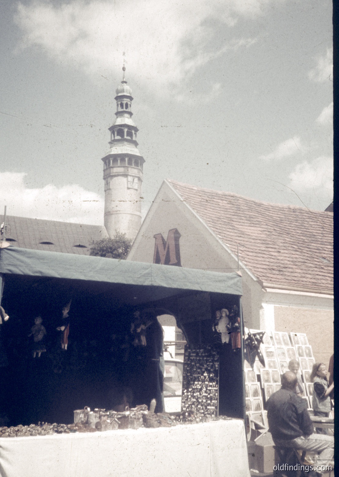 Vintage outdoor market stall with "M" signage under a tiled-roof pavilion, featuring handcrafted wooden toys and folk art. A church tower with a spire and cross looms in the background. Likely Eastern European, mid-20th century.
