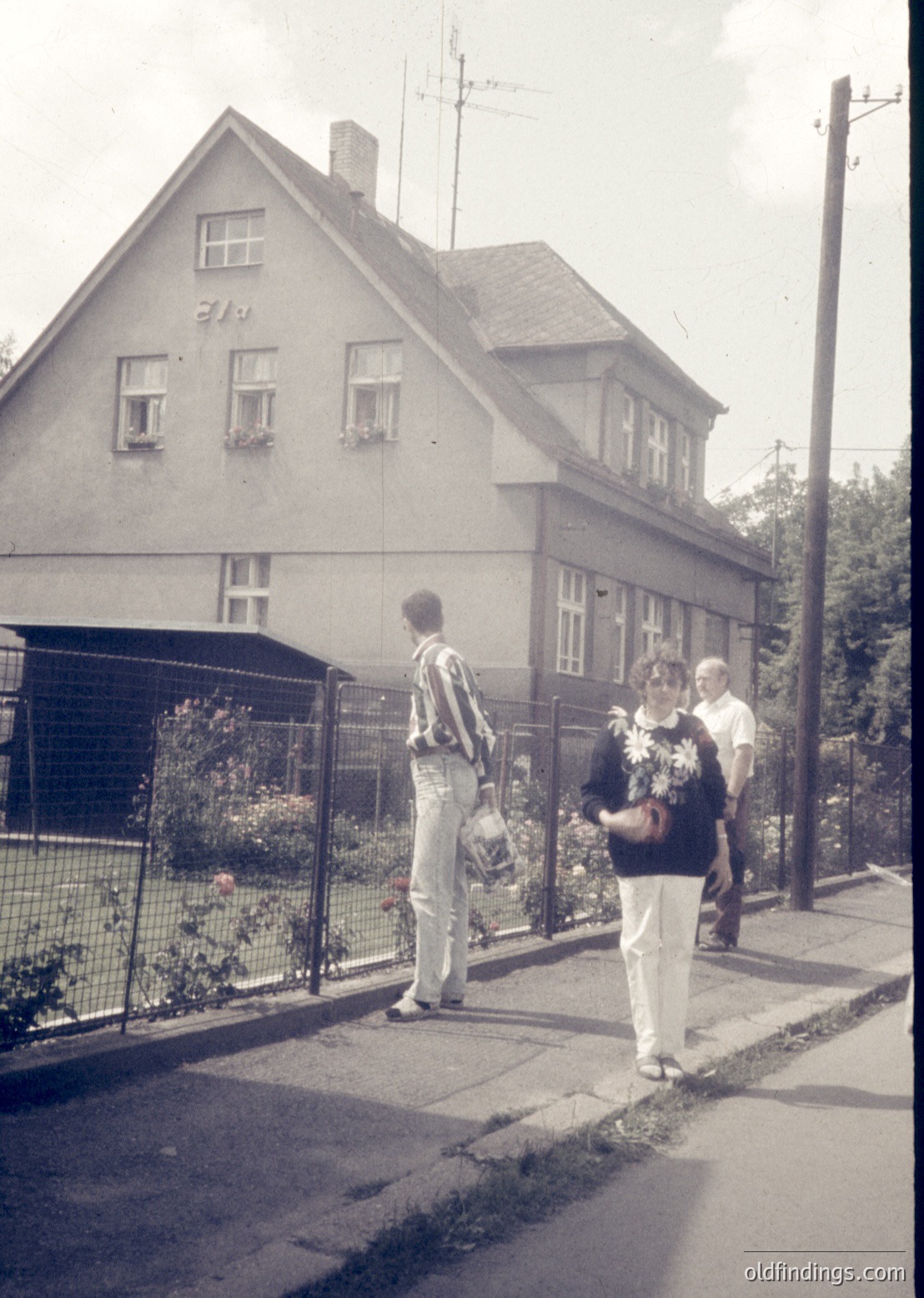 Two men and a woman in 1970s streetwear stand outside a two-story, gabled house with whitewashed walls and wooden-framed windows. The house features a small chimney and a wrought-iron fence. The scene suggests a residential neighborhood. [Mid-century European suburban home with vintage 1970s attire ]