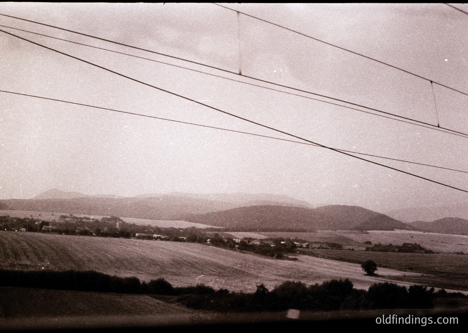Mid-20th century rural landscape with rolling hills and open fields, framed by power lines. Vintage black-and-white grain adds nostalgic texture. Likely Eastern European agricultural region, mid-century farming era.