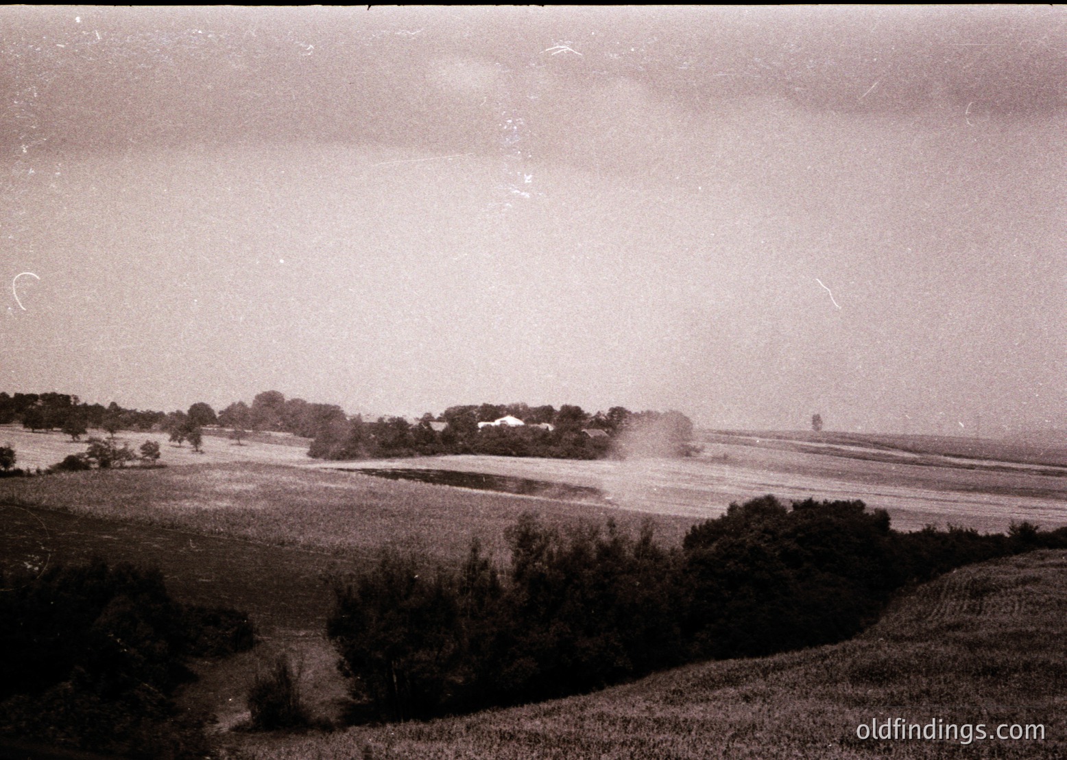 Vintage black-and-white landscape showing rolling farmland with scattered trees and distant structures, likely agricultural. Overcast sky enhances moody, timeless atmosphere. Suggests early-to-mid 20th century rural setting.