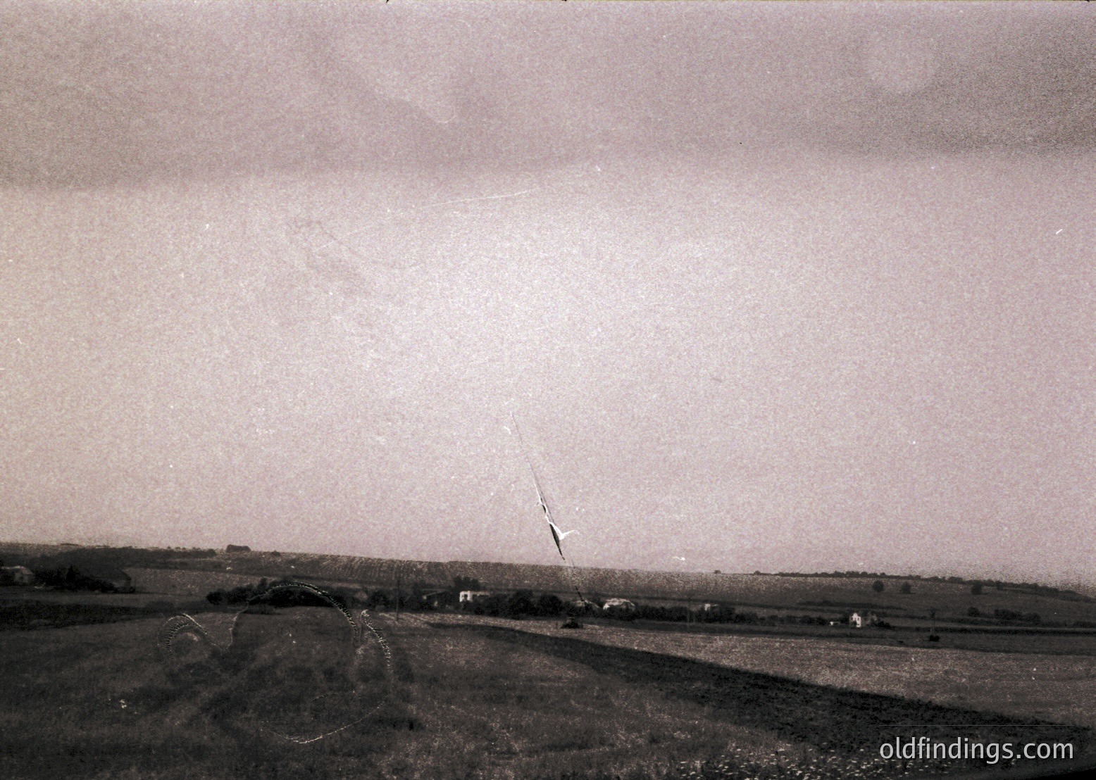 Vintage aerial view of rural landscape with low-lying clouds and rolling terrain. Distinctive diagonal power line cutting through horizon, sparse farmhouses and fields below. Likely mid-20th century agricultural region.